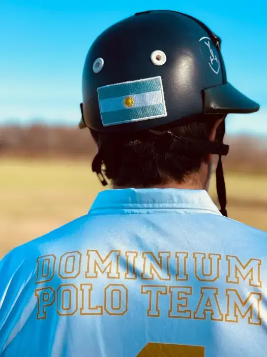 A man from behind wearing a Dominium Polo Team jersey and helmet with an Argentina flag patch, in an open field
