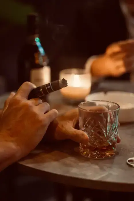 Close-up of a hand holding a Dominium cigar and a glass of whiskey at a table