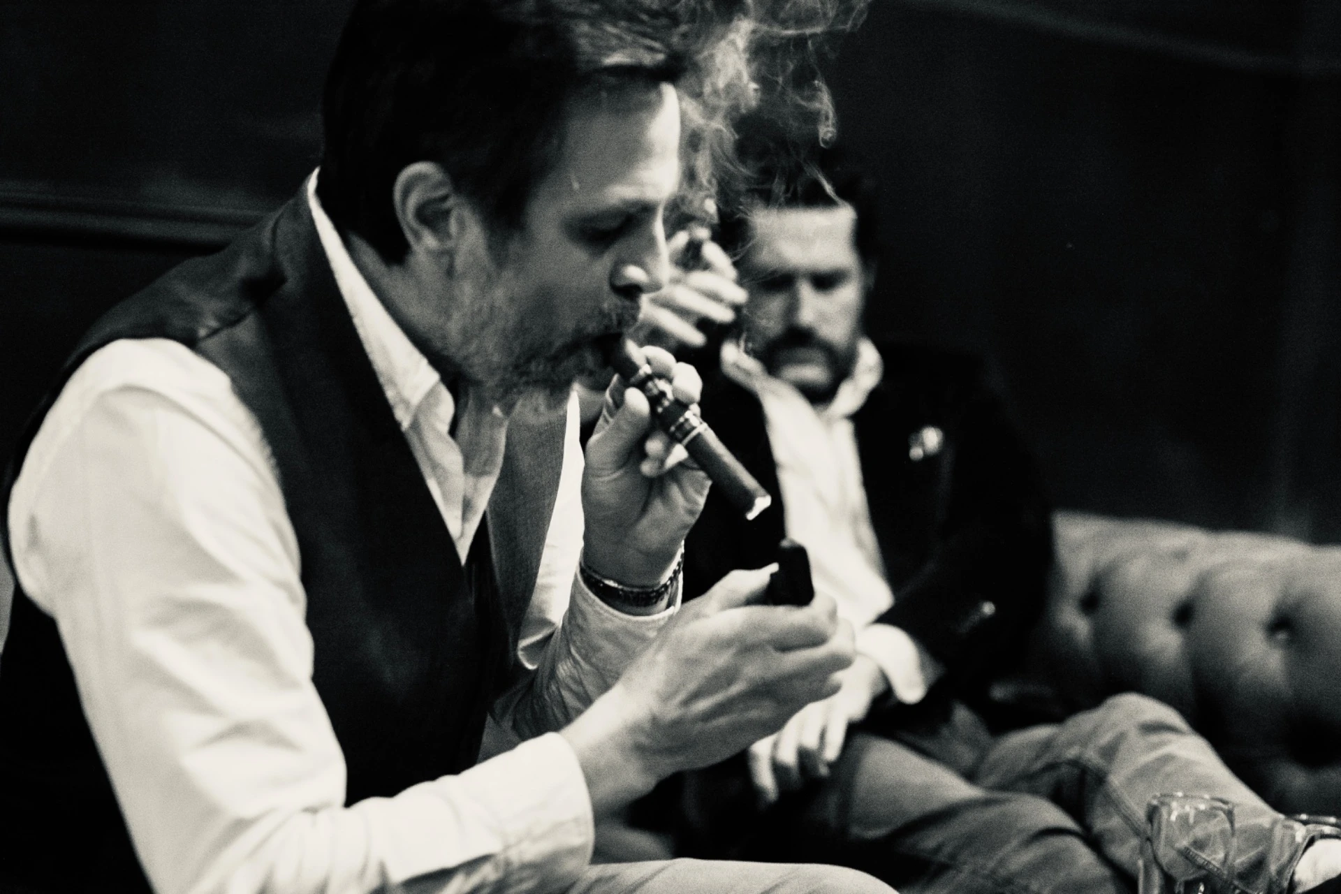 A man lighting a cigar on a leather couch, another man in the background